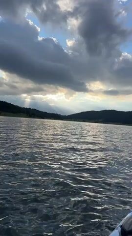 A kayaker is moving across a lake, with the camera focused on the front of the kayak. The sky is partly cloudy with sun rays breaking through. There are hills in the background. Another kayaker is visible in the distance.