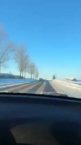 The video shows a car driving on a snowy road on a sunny day. The road is mostly clear, with patches of snow and ice in the tire tracks. The sides of the road are covered in snow, and there are trees on either side. The sky is clear and blue. Another car passes by in the opposite direction. The camera is inside the car, looking out through the windshield. Music plays in the background.