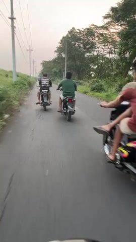 The video shows three young males riding motorcycles down a paved road. The person filming is riding behind the other two, facing away from the camera. The sky is overcast and the sun appears to be setting. The road is lined with vegetation and trees. The motorcycles are all small, dirt bike style. The individuals appear to be teenagers. The scene suggests a rural setting.