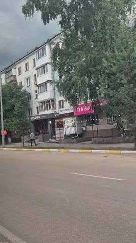 The video captures a street view of a multi-story apartment building with storefronts at the ground level. A few trees and bushes are visible in front of the building. A silver sedan is driving on the street in front of the building. A person is walking on the sidewalk in front of the building. The sky appears overcast.