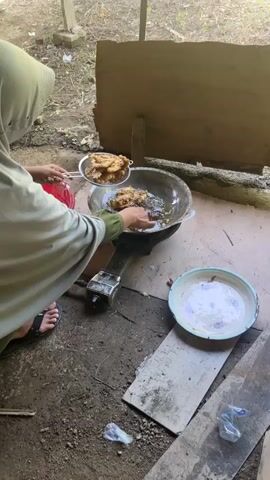 A person wearing a light green hijab and an olive green sleeve is frying food in a wok filled with oil. The wok is on a portable gas stove. The person is using tongs to move pieces of golden-brown fried food from a metal sieve into the wok. Another person's hand, wearing a red sleeve, is visible to the left, holding a red bowl. A white plate with a floral pattern sits on a piece of wood to the right of the wok. The ground is dirt and debris. The background consists of wooden structures and vegetation.