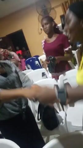 The video depicts a group of people, mostly women, in what appears to be an indoor space. There are white plastic chairs, a fan, and some bags in the background. One woman wearing a yellow shirt and a black and white skirt seems to be dancing and interacting with the camera. Other women are seen in the background, some in maroon shirts and one with a pink strap, holding items in their hands. The overall atmosphere seems celebratory or lively. The camera movement is unsteady and close to the subjects, making it difficult to clearly see the details of the room.