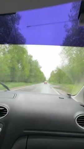 The video shows a view from inside a car driving on a wet road. It is raining. The car is moving forward, following another car in front. The road is surrounded by trees. A blue sun visor is visible at the top of the frame. The interior of the car is visible, including the dashboard, air vents, and a piece of equipment below the air vents.  The windshield wipers are activated.