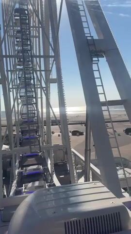 The video is shot from the perspective of someone inside a gondola of a Ferris wheel, looking outwards and upwards. The gondola interior is visible in the foreground, showing a white surface with a gray grate. The massive white metal structure of the Ferris wheel dominates the frame, with its spokes, beams, and ladders extending upwards. In the background, the bright sun is visible through the spokes, creating lens flares and highlighting thin clouds in the clear blue sky. Below the Ferris wheel, a beach with sand and rows of lounge chairs can be seen, extending towards a calm ocean with a visible horizon. There are no people clearly visible in the video, but the beach appears to be empty or sparsely populated. The Ferris wheel itself has multiple gondolas, some with blue lighting visible, ascending along its circular track. The overall impression is a sunny day at a seaside amusement park.