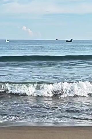 The video shows a serene ocean scene. Gentle waves are rolling onto a sandy beach. The water is a deep blue, and the sky above is partly cloudy with a hazy sun. In the distance, several small boats are visible on the horizon. The overall atmosphere is peaceful and tranquil.