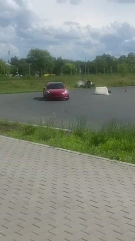A red Tesla Model 3 is seen on a paved track, possibly an autocross course. Orange cones are scattered around the track, indicating a course layout. The car is moving, appearing to execute a maneuver, possibly a drift or a tight turn. In the background, there are grassy fields, trees, and a cloudy sky. There are also some white barriers and tires placed on the track. The car has red registration plates and appears to be in motion. The footage is shot from a slightly elevated position, looking down at the car and the track. The overall impression is of a performance driving event or practice session.
