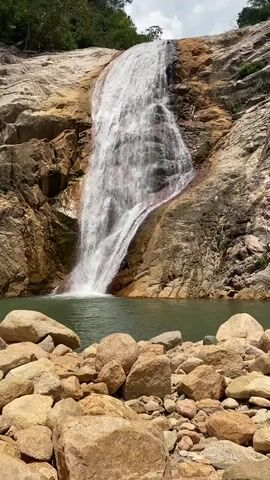 A video shows a waterfall cascading down a rocky cliff into a pool of water. The water is clear and green, and the rocks are varied in color, from light tan to reddish-brown. In the foreground, there are large boulders and smaller stones. A person is visible in the water, swimming. Trees are visible at the top of the cliff. The sky is overcast.