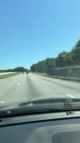 The video captures a first-person view from inside a vehicle traveling on a multi-lane highway under a clear blue sky. The asphalt road is marked with white lines, and there are several other vehicles visible in the distance. To the right of the highway, there is a raised embankment covered with trees and foliage. A blue sign with text and symbols is visible on the right side of the road. The dashboard of the vehicle is visible at the bottom of the frame.