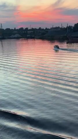 A wide shot shows a sunset over a body of water. The sky is a blend of pink, orange, and blue. The water is calm with gentle ripples. A small white boat is moving across the water from left to right, leaving a wake behind it. In the distance, along the shoreline, there are industrial buildings, cranes, and piles of sand or gravel. The overall scene depicts a tranquil evening on the water with hints of industrial activity in the background.