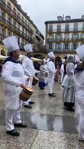 The video depicts a group of people dressed in chef-like attire, including white hats and coats, playing drums in a public square on a cloudy day. The square is surrounded by old European-style buildings with multiple windows and archways. Some individuals wear transparent plastic covers over their outfits. The ground appears wet, likely due to rain. Other people can be seen in the background, some also playing drums, and a few bystanders watching the performance.