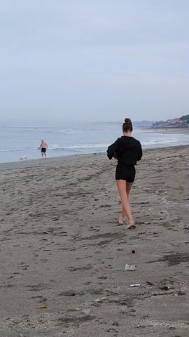 Here's a description of the video:
The video shows a beach scene with a woman walking away from the camera. She has her hair in a bun and is wearing a black hooded jacket and black shorts. She is walking barefoot on the sand. In the background, a man is running along the shoreline with a dog. The beach appears to be somewhat littered with debris. The sky is overcast, and the ocean has gentle waves.