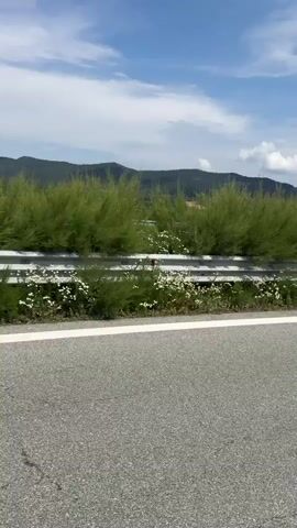 The video is a first-person perspective recording from a vehicle traveling on a highway. The road is paved and has a white line marking the lane. A metal guardrail runs alongside the highway, separating it from the grassy shoulder and vegetation. In the background, there are rolling green hills and mountains under a bright blue sky with scattered white clouds. The scene appears to be a scenic rural or semi-rural landscape. There are no people visible in the video, and the primary action is the forward movement of the vehicle.