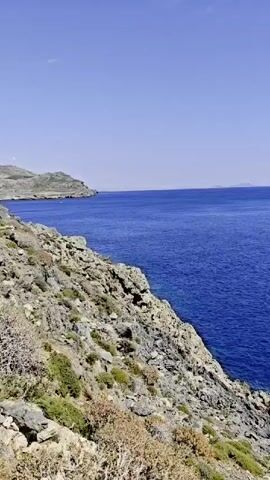 The video shows a view of a blue sea from a rocky coastline. The sky is clear and blue. The coastline is covered in rocks and sparse vegetation. The sea stretches out to the horizon. The camera remains static throughout the video.