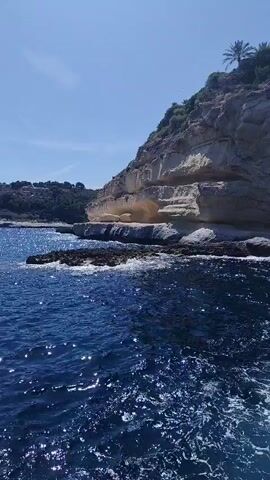 This video showcases a boat ride along a rugged coastline under a clear, sunny sky, suggesting a pleasant day. The camera is positioned on a boat, looking towards a steep, rocky cliff face. The cliffs are a light, sandy color with visible layers and crevices, and are topped with lush green vegetation and a few palm trees. A large, modern white building with a flat roof is situated on the hilltop, overlooking the sea.

The water is a deep, vibrant blue and appears choppy, with whitecaps indicating movement. The boat has metal railings and steps visible in the foreground, hinting at it being a tour or passenger vessel. The activity is a scenic boat tour, likely exploring coastal caves or points of interest. The time of day appears to be daytime, given the bright sunlight and clear blue sky. The location cues, such as the rocky cliffs, Mediterranean vegetation, and the style of the building, align with the provided context of Sol De Mallorca, Spain. No text is visible in the video.