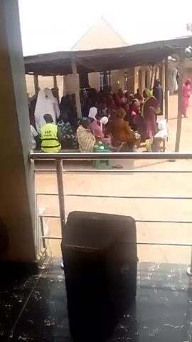 The video shows a group of people gathered outdoors under a covered area. They are seated on chairs and mats. Some people are wearing traditional clothing. One person is wearing a yellow vest with the letters "YAF" on it. A black speaker is in the foreground. The view is from inside a building looking out onto the gathering. The building has a tiled floor and a metal railing. The weather appears sunny.
