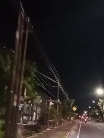 The video shows a nighttime street scene. There are power lines overhead. A house with a blue facade and a covered patio is on the left. A flagpole with the Indonesian flag is visible in front of the house. A yellow diamond-shaped sign with an exclamation mark is on the right side of the road. Further down the road, streetlights and car headlights are visible. Trees and foliage are present on both sides of the street. The ground is paved and appears to be a sidewalk and a road. The sky is dark and clear.