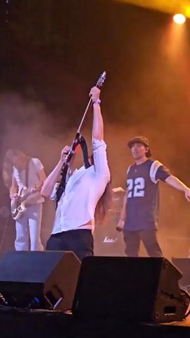 The video shows a live musical performance with three individuals on stage. The central figure, a long-haired person wearing a white collared shirt and dark pants, is energetically playing a black electric guitar, holding it above their head and then bending down. To the left, another long-haired individual in a white t-shirt and light-colored pants plays a white electric guitar. To the right, a person wearing a dark baseball cap, a blue and white athletic jersey with the number 22, and dark pants stands holding a microphone, gesturing with their right hand. The stage is filled with smoke or fog, illuminated by stage lighting that shifts between yellow and red hues. Large black speaker cabinets are visible in the foreground. Marshall amplifier logos are visible behind the guitarist on the left.