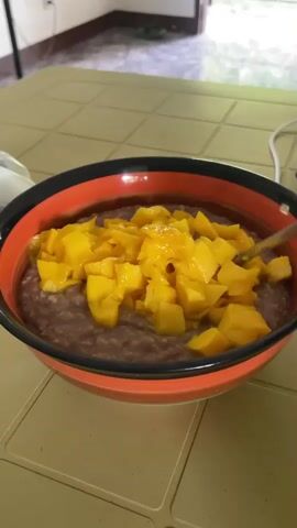 A close-up, eye-level shot shows a bowl of what appears to be purple porridge topped with diced yellow fruit, possibly mango. The bowl has a black and orange rim. A metal spoon is being used to scoop a portion of the porridge and fruit from the bowl. The bowl is sitting on a table with a tiled surface. In the background, a white cord is visible and a doorway leads into another room. The lighting is bright and appears to be natural light from a window.