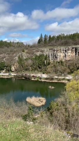 The video pans across a scenic view of a lake surrounded by natural landscapes. Initially, the camera overlooks a dark green lake with small patches of vegetation in it. A winding dirt path borders the lake, alongside a road with parked cars. In the background, a large cliff face rises, crowned with a dense forest. The camera then pans to the right, revealing a grassy dam-like structure with a group of about ten people standing on it. Continuing the pan, the video reveals a large white house with a red roof, set atop the cliff overlooking the lake. The camera continues panning right to show more of the lake and the surrounding forest. The sky is partly cloudy with patches of blue.