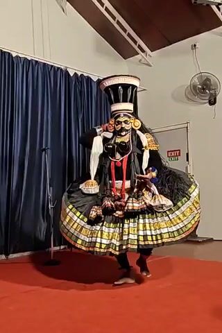 The video shows a person in a elaborate costume performing a traditional dance. The dancer, who appears to be male, is dressed in a black and gold plaid skirt-like garment with a tall, ornate headpiece. He is wearing a black mask with golden accents and has long, dark hair. The dancer moves rhythmically on a red-carpeted stage, making gestures with his hands. In the background, a man in traditional Indian attire plays an instrument. The room has blue curtains, a white ceiling with wooden beams, and an exit sign on a white door. A microphone stand is visible near the front of the stage.