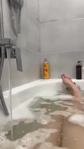 The video shows a person in a bathtub filled with water and bubbles. The person's legs and feet are visible, but the rest of their body is submerged or out of frame. A faucet is running, adding more water to the tub.  On the edge of the tub are several bottles of bath products. The bathroom has light gray tiled walls.