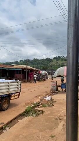 A small truck loaded with white sacks is parked on the left side of a dirt road. Several market stalls with awnings line the right side of the road. Two people are visible walking between the stalls, and a motorcycle is parked in front of one of them. A hilly, green landscape is visible in the background under a cloudy sky.