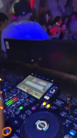 The video shows a DJ performing in a crowded nightclub with bright, colorful lights. The DJ is using a Pioneer DJ controller with a touchscreen interface. The dance floor is full of people dancing and enjoying themselves, illuminated by blue, pink, green, and red lights. Some people are holding drinks, including a bottle of Corona. The environment appears lively and energetic.
