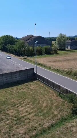 A white convertible car drives down a two-lane asphalt road bordered by a low concrete wall and a metal fence. Beyond the road, fields of dry grass and scattered trees are visible, along with several rural buildings under a clear blue sky. A second white car, a hatchback, approaches from the opposite direction and passes the convertible.