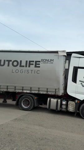 The video displays a white FAW J7 heavy-duty truck, recognizable by the "FAW" logo on the cab's roof and near the side mirror, along with a stylized "J7" graphic on its side. The truck's cab is white with tinted windows and large side mirrors, and silver fuel tanks are visible below. It is coupled with a white curtain-sided trailer.

The trailer features the text "AUTOLIFE LOGISTIC" prominently on its side, along with a "BONUM MAIN LINE" logo above it, indicating a transportation or logistics service.

The vehicles are parked on a gravel or dirt ground. The background includes a building with a dark roof, a red fence, and some distant foliage, all under an overcast sky. The video provides a static view, initially showing the full side profile of the truck and trailer, then zooming in to highlight the details of the truck's cab. No people or active events are visible. The setting is Kamyennoye, Russia.