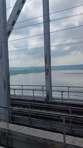 A train crosses a bridge over a river. The bridge is made of white metal girders. The river is wide and calm, with trees on the far bank. The sky is cloudy. The train tracks are visible in the foreground.