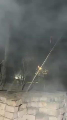 The video shows a fireworks display at night. A dark wall is visible in the foreground. Fireworks explode in the sky, creating colorful bursts of light. Smoke fills the air. The camera is pointed upward. Bright, golden fireworks fill the sky, followed by smaller bursts of red and white. The sound of the explosions can be heard. The scene is festive and celebratory.