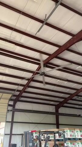 A large industrial ceiling fan with multiple blades spins rapidly in a warehouse. The fan is suspended from the ceiling, which is made of white corrugated metal and supported by dark red steel beams. In the background, there is a forklift and stacked boxes. The overall impression is that of a functional warehouse environment.