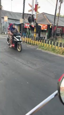 The video shows a person riding a scooter on a road. The person is wearing a headscarf and a patterned dress. They are carrying a red backpack on their back. The scooter is black and has gold accents. The road appears to be paved and is in a rural or semi-urban setting. In the background, there is a railroad crossing with a signal light and a barrier. There are also buildings visible, one of which has a sign with red and yellow lettering. The lighting suggests it is daytime, possibly late afternoon or early morning. The video is shot from the perspective of another person on a scooter, with the handlebars and a mirror of that scooter visible in the foreground.