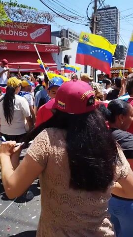 Here's a detailed description of the video:

The video shows a vibrant street celebration. A crowd of people are gathered, waving Venezuelan flags. The person filming is in the middle of the crowd, turning around. Many of the people in the crowd are wearing hats, some with the Venezuelan flag. There's a "Minimarket Alonso" with "Coca-Cola" signs visible in the background. Several high-rise buildings can be seen in the distance. Some people are drinking from cans. The overall atmosphere seems lively and celebratory.