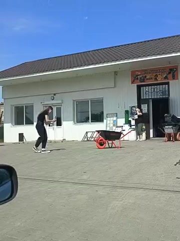 A video shows a person in black clothing standing on a paved area outside a white building with a brown roof. The building has several windows and a large open doorway with a sign above it displaying tools. A black and red wheelbarrow is positioned in front of the building. Another person in green pants and a dark top stands near the doorway. The person in black is holding a phone and appears to be recording or taking pictures. The sky is blue with a few wispy clouds. The video is shot from inside a vehicle, with the car's frame visible in the lower left corner.