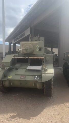 The video shows a line of vintage military tanks parked under a covered pavilion labeled "Veterans Pavilion". The tanks are painted in various shades of green and brown and have military markings and numbers on them. They are lined up on a dirt and gravel surface under the shelter of the pavilion. The camera pans from left to right showing the tanks. The scene is outdoors on a bright, sunny day. The tanks appear to be on display.