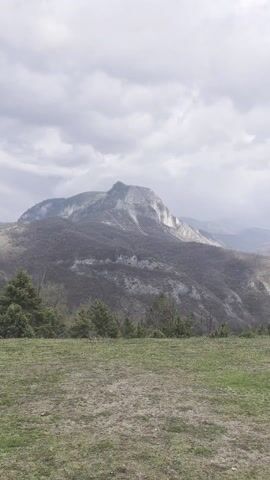 The video features a slow, sweeping panoramic shot of a mountainous landscape, likely in the vicinity of Laki, Bulgaria. The camera pans from left to right, revealing a vast, rugged natural environment under a dynamic, cloudy sky.

In the foreground, a gentle slope covered in patchy grass, showing both greener and drier sections, occupies the lower third of the frame. As the view progresses, a circular stone fire pit becomes visible on the right side of this grassy area, along with a small portion of a building with a dark red-tiled roof, suggesting an established overlook or recreational spot.

The midground is dominated by steep mountain slopes, heavily forested with a mix of bare deciduous trees and scattered dark green evergreens, indicating early spring or late winter. Valleys and ravines cut through the terrain, adding to its ruggedness.

In the background, multiple layers of mountain ranges recede into the distance. On the initial left side, a prominent, craggy peak with sparse vegetation stands out, catching some diffused light. Further mountain ranges appear progressively hazier, blending with the atmospheric conditions.

The sky is largely overcast with varying shades of grey and white clouds, but patches of brighter sky and distinct white cumulus clouds are visible, suggesting a changeable weather pattern or breaks in the cloud cover. The lighting is diffused, consistent with an overcast daytime.

No people or visible text are present in the video. The overall scene evokes a sense of expansive natural beauty and tranquility within a wilderness setting.