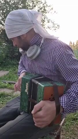 A man is playing a green and brown "HOHHA" harmonica outdoors. He is wearing a purple and white plaid shirt, gray pants, and a white head covering, and he has gray headphones around his neck. He is sitting down and looking at the instrument as he plays it. The background consists of green grass and trees. The lighting suggests it is near dusk.