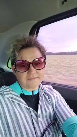 A woman is sitting in the passenger seat of a car, looking at the camera. She is wearing large, oval-shaped sunglasses with a burgundy frame, a striped shirt with white and grey stripes, and a teal-colored undershirt. Her hair is short and styled. The window next to her is open, and a blurry, light-colored field can be seen outside. The sky is a hazy purple-pink color. The woman is smiling.