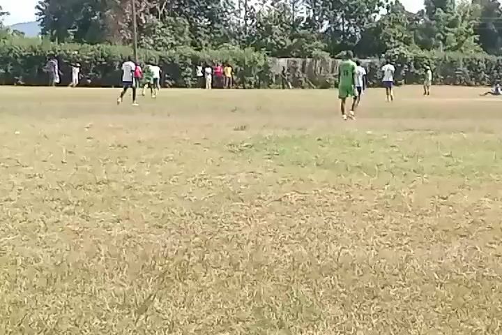 The video shows a soccer game being played on a grass field.  Several players wearing green and white uniforms are scattered across the field. Some players are running, while one appears to be on the ground. Other people, potentially spectators, stand along the sidelines in the background.  Trees and utility poles can be seen in the background.  The grass appears to be dry in places, and the lighting suggests a daytime setting.