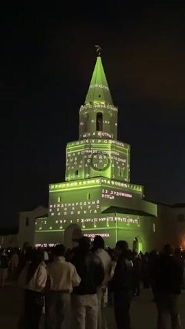 At night, a crowd of people stand facing a tall, multi-tiered stone tower. The tower has a green pointed spire topped with a weather vane. Projections of green text and imagery appear on the tower. The projections change to a pattern of white lines that resemble scribbles. The projections then change to bright colors with images of a large eye and abstract shapes. The tower is part of a larger building complex. The sky is dark.