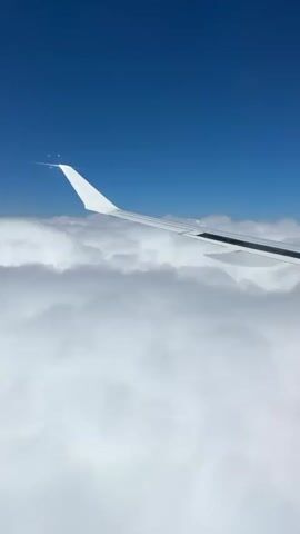 The video shows a view from an airplane window, looking out at a vast expanse of white clouds under a clear blue sky. The wing of the airplane is visible in the foreground, extending across the frame. There are small, bright objects on the tip of the wing, possibly ice crystals or frost. The camera angle suggests the airplane is in flight, moving through the sky. The overall impression is one of serene aerial travel.