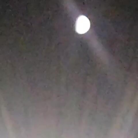 The video displays a hazy, slightly out-of-focus view of a waxing gibbous moon in a dark sky. A beam of light, possibly from a projector or spotlight, is visible, extending from the bottom of the frame towards the moon, with the moon appearing to be partially within this light beam. The overall image quality is low, with visible digital noise and a soft focus. There are no people or distinct objects identifiable in the scene other than the celestial body and the light beam.