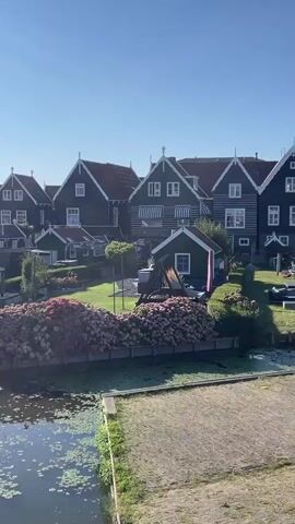 The video shows a picturesque waterside scene with traditional dark-colored houses featuring white trim and peaked roofs. In the foreground, a calm body of water is visible with lily pads and green algae. Along the water's edge, there are wooden posts and a cobblestone path. Behind the water, a lush green lawn is dotted with trees and shrubs. Dominating the middle ground is a vibrant display of pink and purple hydrangeas. A small, dark green shed with white trim and a window stands on the lawn, next to a wooden swing set. Further back, several larger houses are arranged in a row, their facades adorned with white-framed windows and striped awnings. Outdoor furniture, including a dark sectional sofa, is visible on the lawn. The background is a clear blue sky.