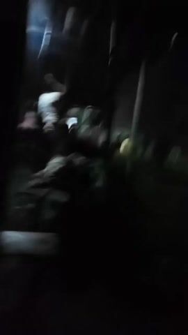 The video shows a dark, outdoor scene at night. A light source briefly illuminates the area, revealing several people gathered on what appears to be a bridge or elevated structure. Some individuals are visible below the structure, possibly in water or a flooded area. The scene is chaotic, with people moving around and some appearing to be in distress. The lighting is poor, making it difficult to discern specific details about the individuals or their actions.