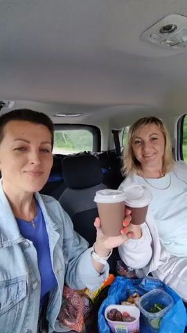 Two women are sitting in the back seat of a car. The woman on the left has short, dark hair and is wearing a blue shirt and a denim jacket. She is holding up two cups of what appears to be chocolate-colored liquid. The woman on the right has blonde hair and is wearing a light-colored shirt. She is smiling and also holding up a cup. In front of them, on the seat, is a blue bag filled with food items, including containers of fruit and vegetables, and a package of meat. The interior of the car appears to be a typical SUV. Outside the car, trees and foliage are visible through the windows, suggesting they are on a road trip or in a rural area. The lighting suggests it is daytime.