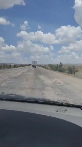 A first-person perspective video from inside a vehicle shows a long, two-lane road stretching towards the horizon under a bright blue sky dotted with fluffy white clouds. The road surface appears worn and cracked in places. On the right side of the road, sparse, dry vegetation and bushes grow in a barren landscape. In the distance, a white truck is visible driving in the same direction as the camera. The sun is shining, casting light on the scene. The interior of the car, including the dashboard and windshield wipers, is visible in the foreground.