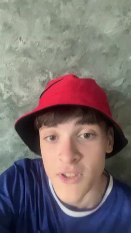 A young person wearing a red bucket hat and a blue shirt is looking directly at the camera with their mouth open. The background is a textured grey wall.