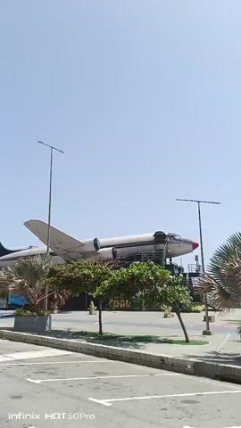 The video captures an exterior view of a unique establishment in Macuto, Venezuela. The main subject is a decommissioned airplane, likely a passenger jet, that has been repurposed as a restaurant or entertainment venue. The plane is situated on the ground, elevated on a dark-colored structure that houses storefronts and seating areas. 

The scene is set outdoors on a bright, sunny day, indicated by the clear blue sky and the strong shadows cast by the airplane and surrounding elements. A tall lamppost stands prominently near the front of the plane. Lush green trees and potted plants add to the environment. The ground is paved, with distinct yellow and white lines painted on it, suggesting it might be a parking area or walkway.

Several storefronts are visible beneath the aircraft, adorned with colorful graphics and signage. One prominent sign reads "Venezuela," and another, partially visible, appears to say "LaParada." Text on one of the storefront windows is blurry but seems to include logos and text in Spanish. The overall impression is a lively, perhaps casual, commercial space utilizing an unusual architectural element. The time of day is clearly daytime due to the bright sunlight. No people are clearly visible in the short clip, but the presence of the establishments implies activity. The mood is likely cheerful and inviting, given the bright weather and vibrant visuals.