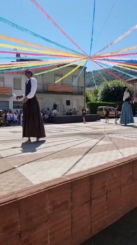 The video captures an outdoor event taking place in a town square. A tall figure, dressed in traditional attire with a long, dark skirt and a vest over a white shirt, stands prominently in the foreground. The figure appears to be a "gegant," a giant figure commonly seen in Catalan festivals. The figure is holding a white object, possibly a scroll or a flag.

In the background, several other figures, some appearing to be children, are present. There is a crowd of onlookers gathered, some standing and others seated. A stage or raised platform is visible, with individuals standing on it. Decorative streamers in various bright colors, including red, yellow, blue, and green, are strung across the sky, creating a festive atmosphere. Buildings with balconies and windows line the square, and one building displays a flag that appears to be the Catalan flag.

The time of day is clearly daytime, with bright sunlight casting distinct shadows on the checkered pavement of the square. The weather appears to be clear and sunny, indicated by the blue sky. The overall mood seems to be celebratory and communal, suggesting a local festival or cultural event. The presence of the gegant, the colorful decorations, and the gathering of people all contribute to this impression. There is no visible text in the video.
